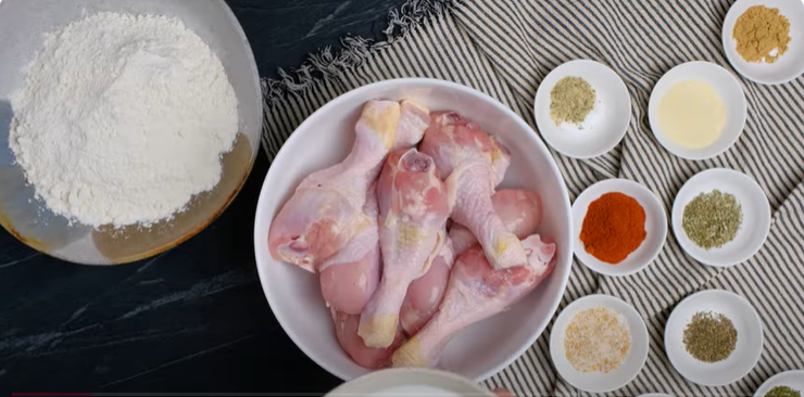 chicken drumsticks in a bowl next to small white bowls with different spices and a bowl of flour. 