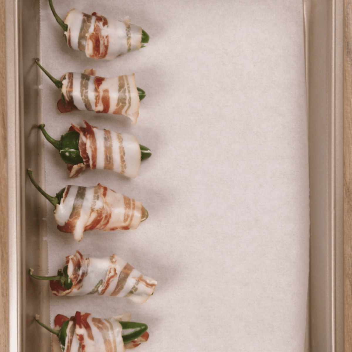 six stuffed bacon wrapped jalapeno poppers in a baking pan with parchment on the bottom.
