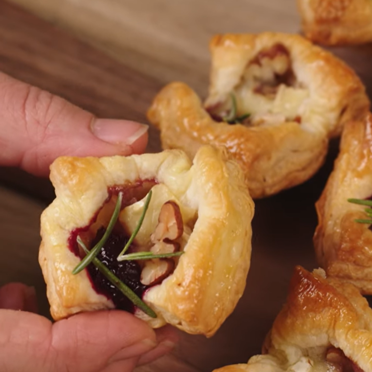 A close-up of a hand holding a golden brown cranberry brie bite made with flaky puff pastry, filled with melted brie cheese, tart cranberry sauce, and chopped pecans, and garnished with fresh rosemary sprigs. More cranberry brie bites are visible in the background on a wooden serving board.