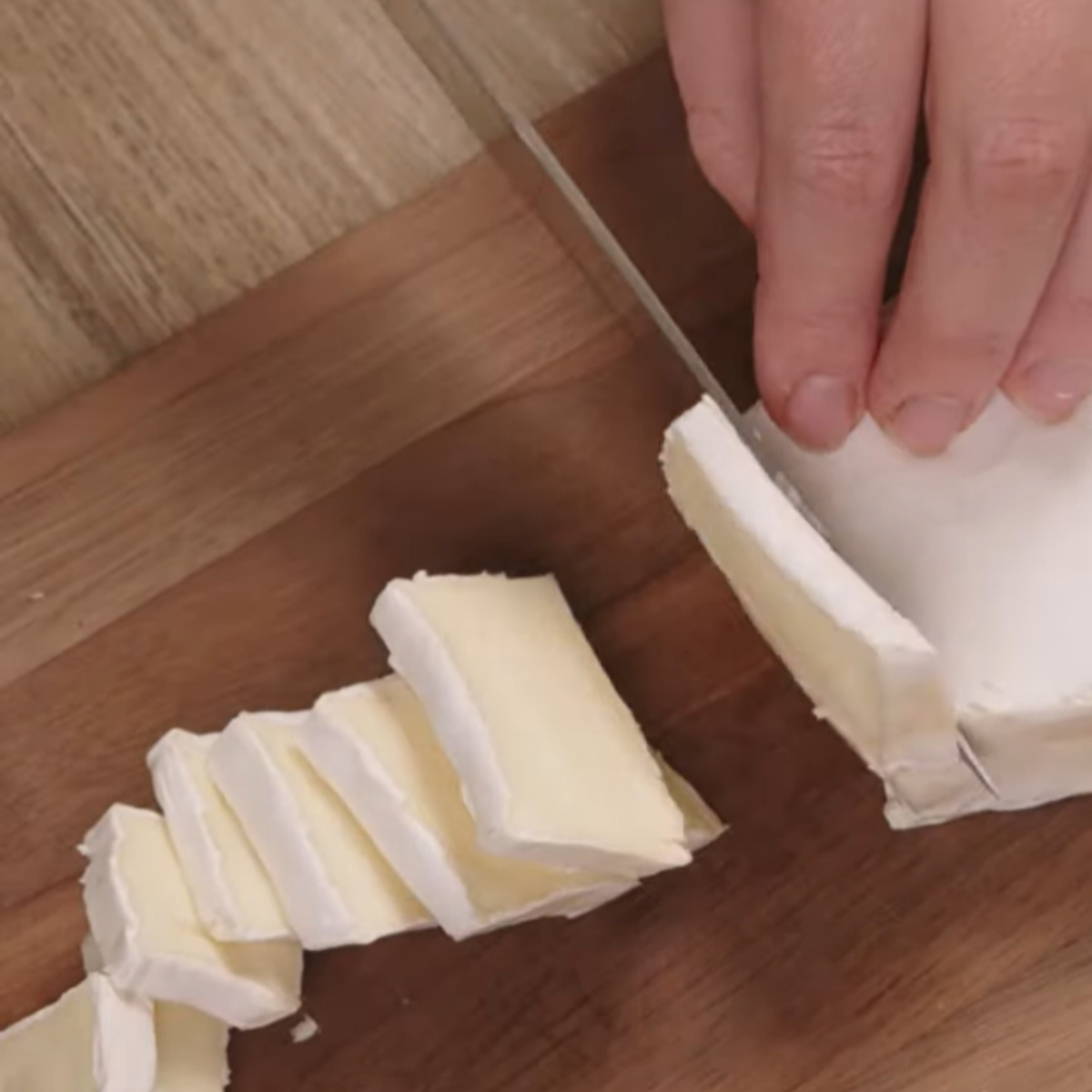 Hands using a knife to cut a wedge of brie cheese into smaller pieces on a wooden cutting board.