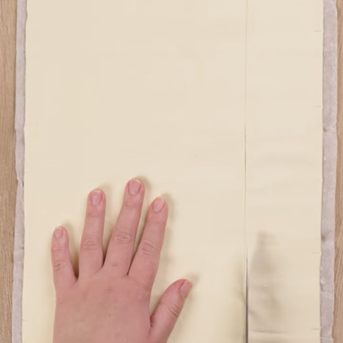 A person's hand using a knife to cut a sheet of puff pastry dough on a wooden surface.