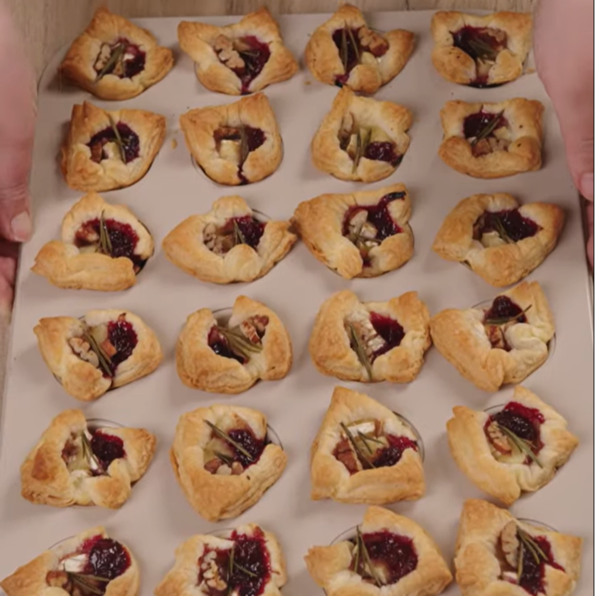 A tray of baked cranberry brie bites, each with a golden brown puff pastry shell filled with melted brie and cranberry sauce, topped with chopped pecans.