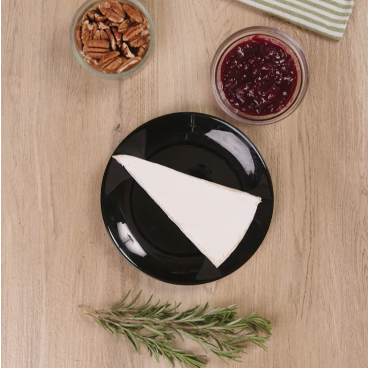 A wedge of brie cheese on a black plate, with a bowl of cranberry sauce and a small dish of pecans alongside.
