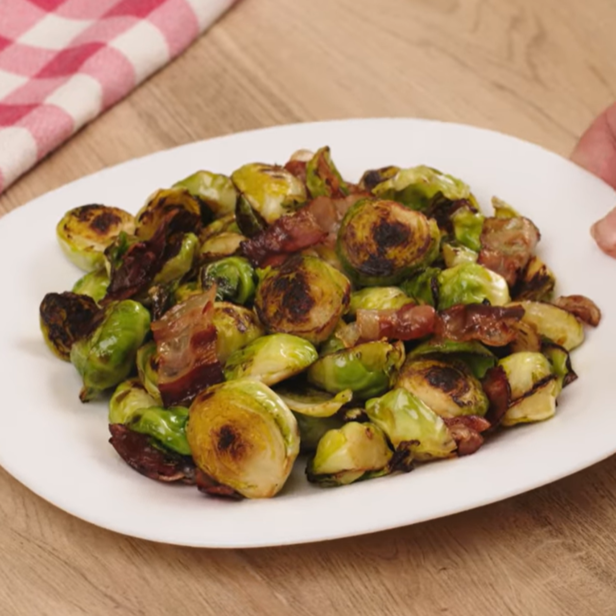 A white plate filled with roasted Brussels sprouts and crispy bacon pieces, caramelized to a golden brown. The dish is placed on a wooden table with a red and white checkered napkin in the background. A hand is partially visible, presenting the plate.