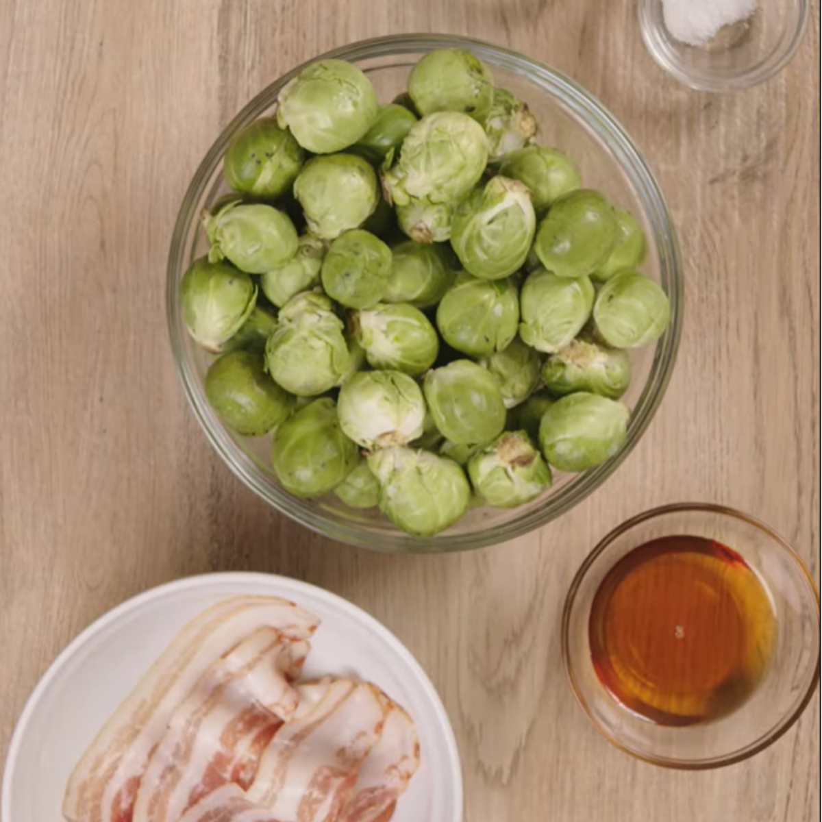 A top-down view of fresh ingredients for maple bacon Brussels sprouts. A glass bowl filled with raw Brussels sprouts sits on a wooden surface, surrounded by small bowls containing salt, maple syrup, and slices of raw bacon on a white plate.