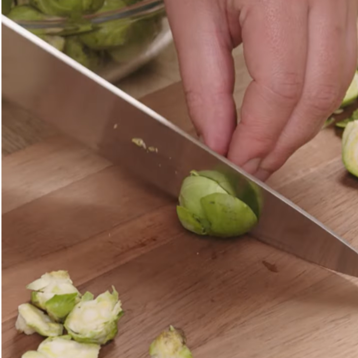 A close-up of a hand using a sharp knife to trim and cut a Brussels sprout in half on a wooden cutting board. Small pieces of trimmed leaves and other Brussels sprouts are visible in the background.